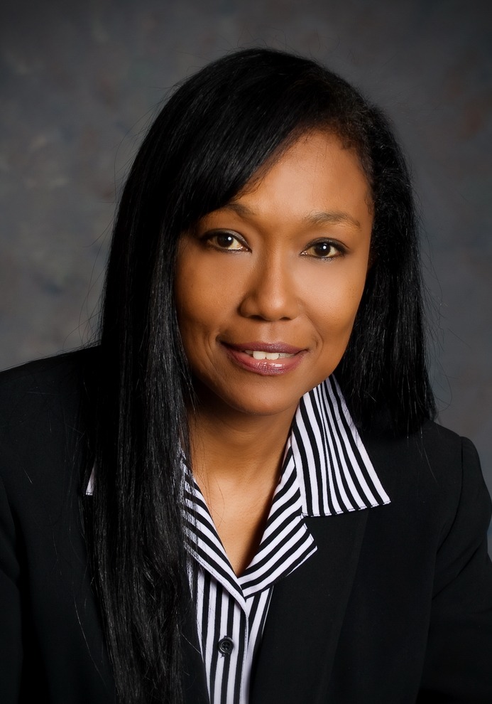 Professional headshot of a woman with long black hair wearing a black blazer over a striped collared shirt. The background is softly blurred.